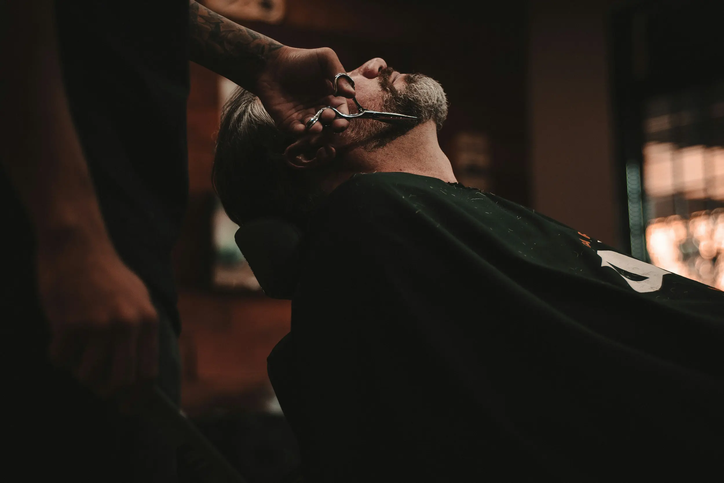 A barber shaping a client's beard with scissors as he reclines in the chair
